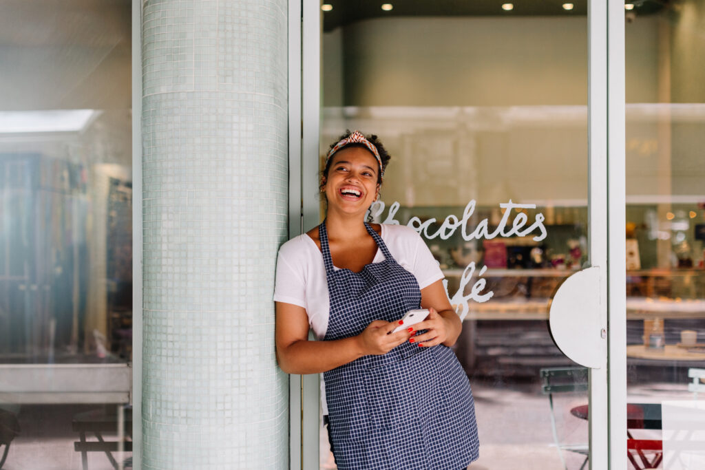 Successful cafe owner, a young woman, stands in her restaurant using a smartphone.