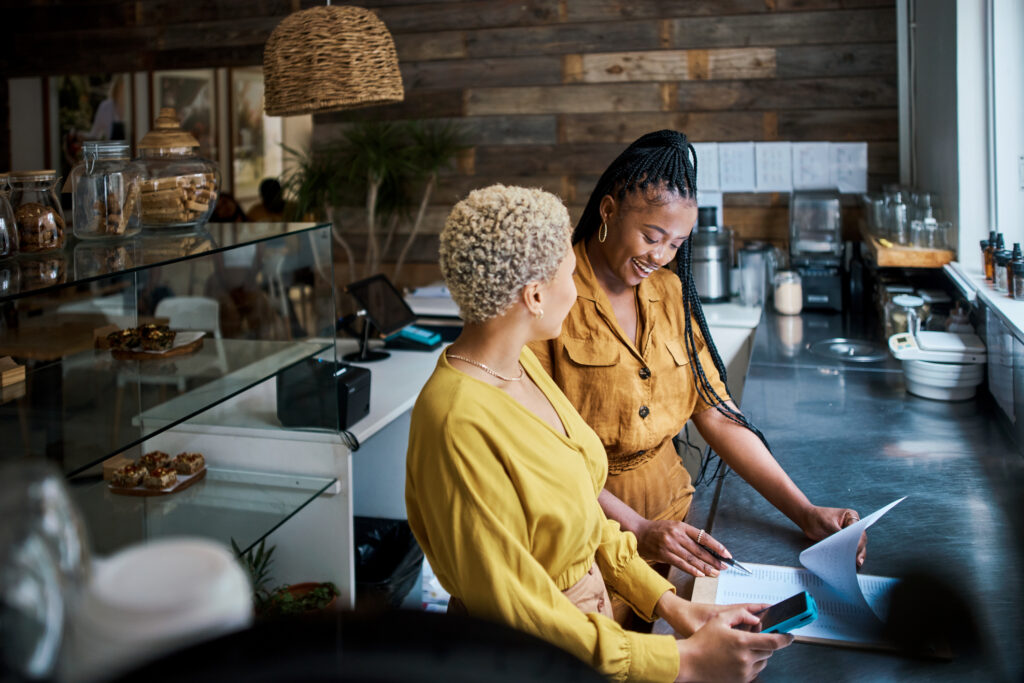 Small business owner training new employee on the job at a coffee shop. Black female entrepreneurs in a partnership collaborating and planning finance and growth together inside the cafe