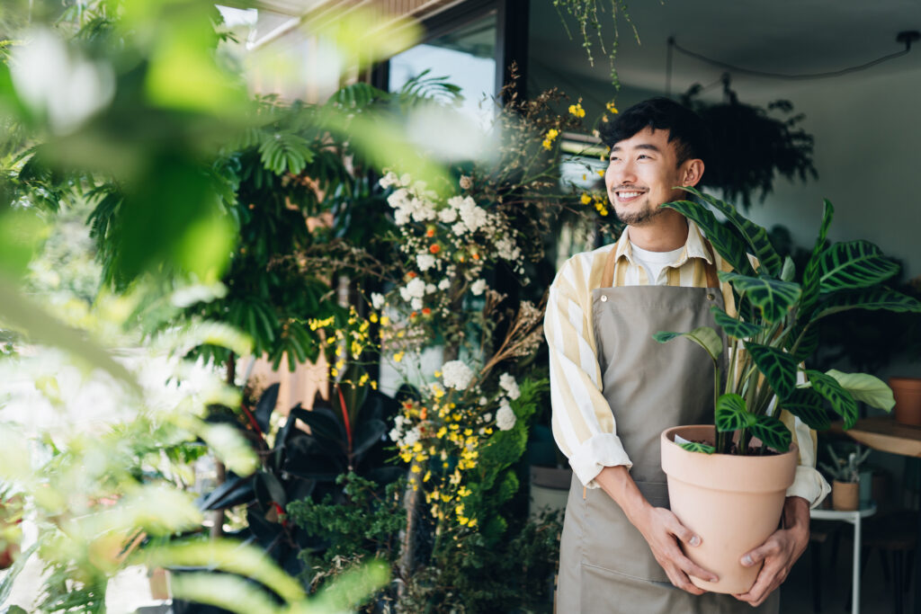 Confident young Asian male florist, owner of small business flower shop. Holding potted plant outside his workplace. He is looking away with smile. Enjoying his job to be with the flowers. Small business concept