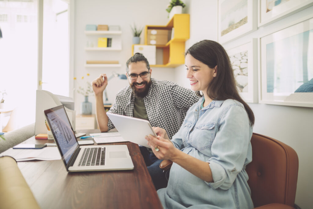 pregnant woman is in front of a laptop and a coworker is showing her paperwork