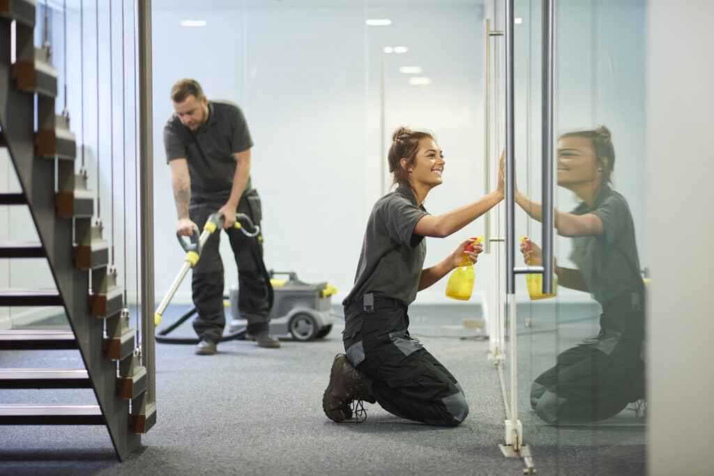 a female cleaning contractor is polishing the glass partition offices whilst In the background a male colleague steam cleans the carpet in a empty office
