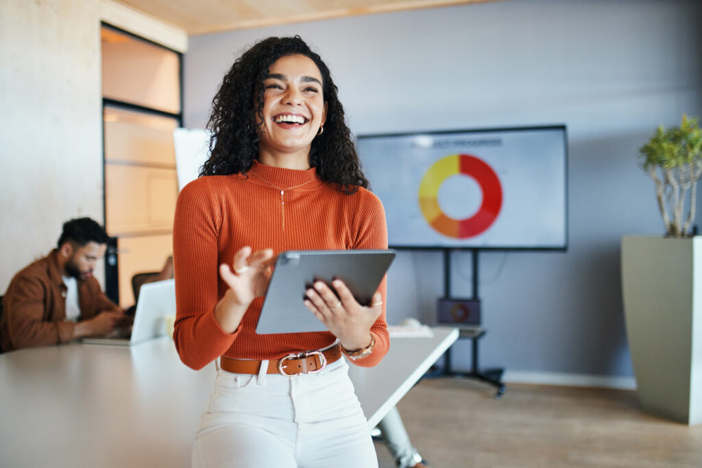Smiling businesswoman with curly hair presents data displayed on a screen behind her in a collaborative office environment.