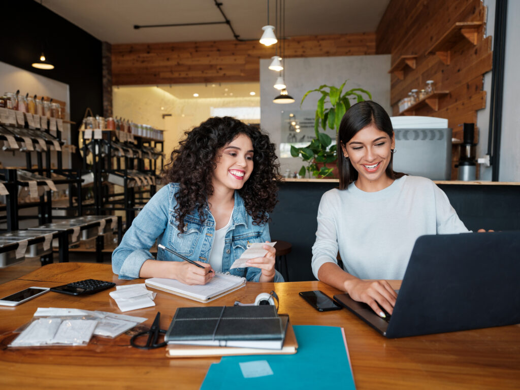 Two young women sitting at a table in an eco-friendly store, using a laptop and smiling.