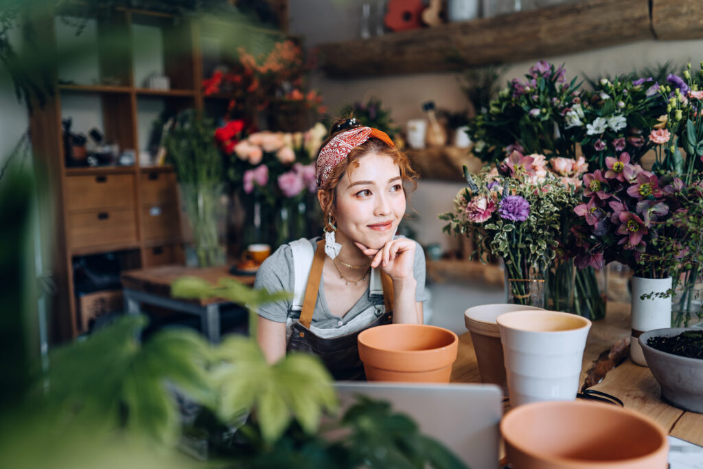 Young female florist, owner of small business flower shop, working on laptop over counter against flowers and plants.