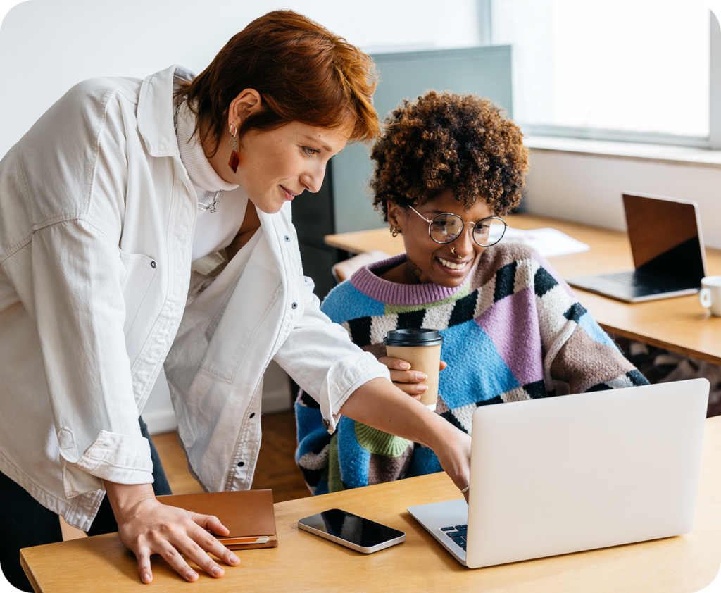 woman looking at her laptop as another woman is standing and pointing to something on the screen