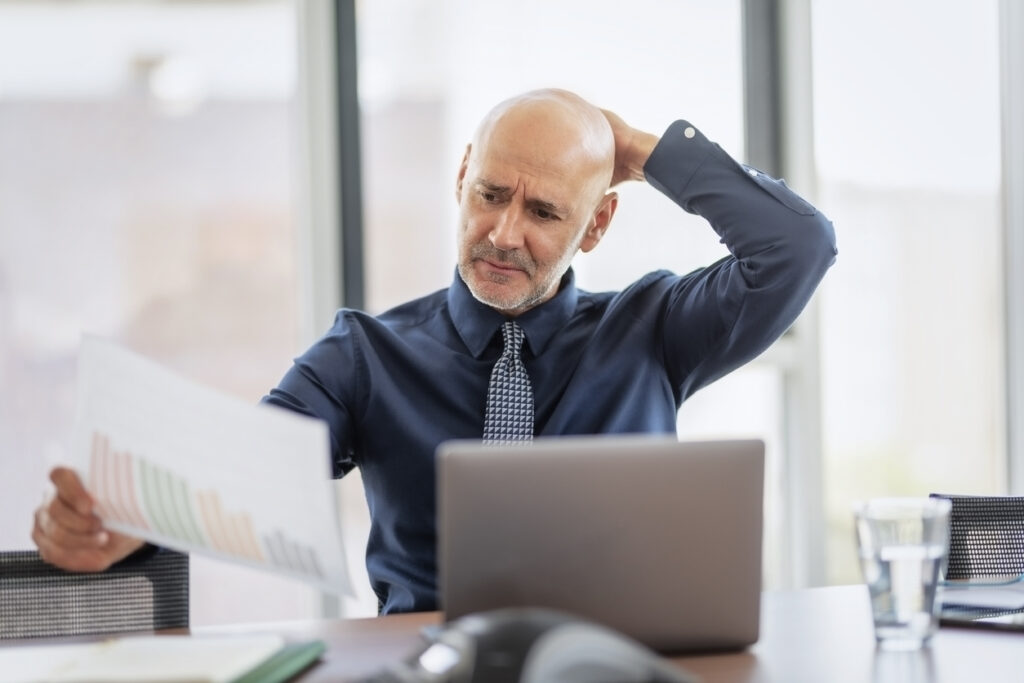 Business owner sitting behind his laptop and holding a paper in his hand while working at the office.