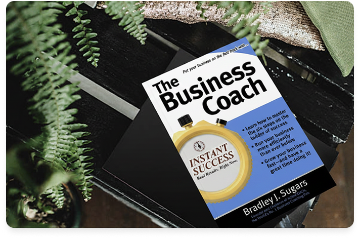 Image of the book The Business Coach on a rustic table painted black, surrounded by houseplants and ferns.