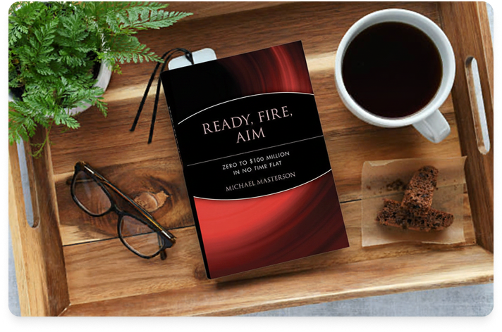 Overhead image of the book Ready Fire Aim on a wood tray with a pair of glasses on the left, a coffee on the right, and a fern in the upper left corner.