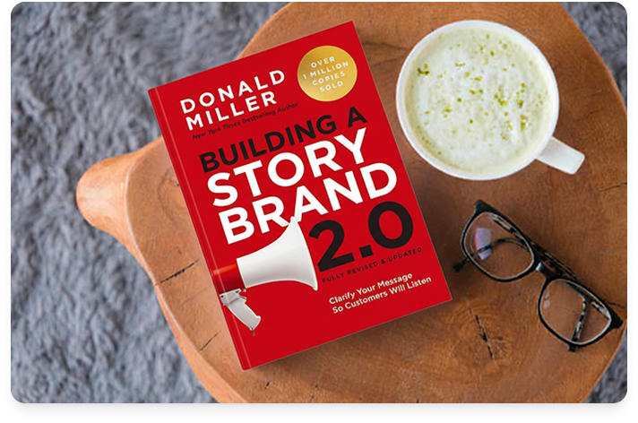 Image of the book Building a StoryBrand on a wood tray with glasses in the lower right corner and a latte in the upper right corner. The tray is on a gray marble table.