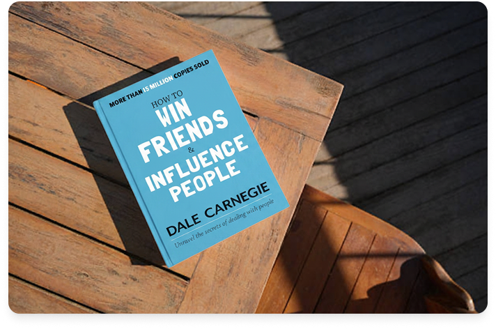 Overhead image of the book titled How to Win Friends and Influence People on a corner of a wood table in an outdoor setting.