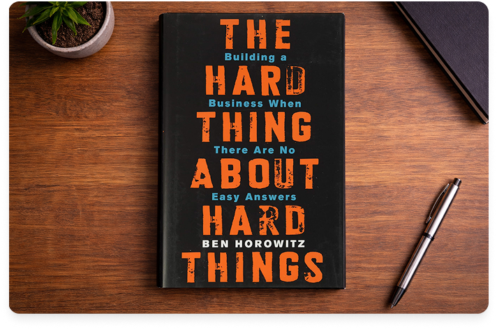The book titled The Hard Thing About Hard Things laying on a reddish wood desk with a pen and a cup of coffee next to it.