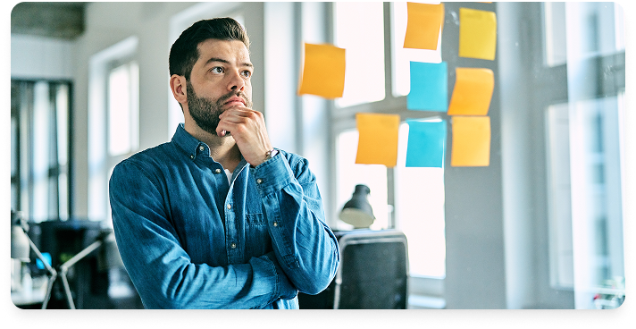 Business man with beard wearing blue shirt in an office, staring at a glass wall with post-it notes, as if assessing ideas from a brainstorming session.