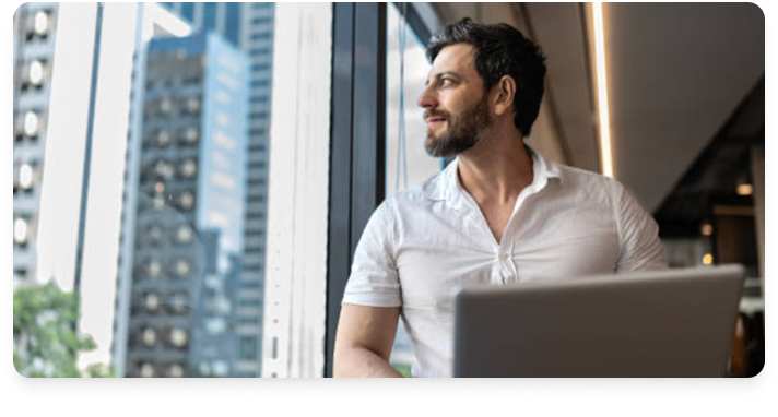 Photo of a man with a beard sitting next to a window. He's staring out the window at skyscrapers, and there's a laptop in front of him.