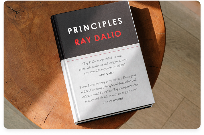 An image of the book Principles by Ray Dalio laying on the edge of a brown desk with a shaft of sunlight across the book cover.