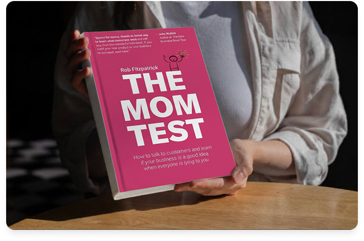 Image of a woman holding the book The Mom Test. The woman is only partially visible, but you can see her hand holding the book on a desk.