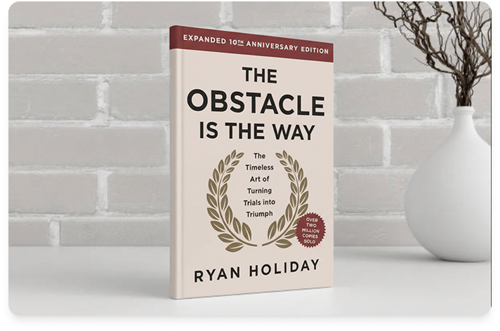 Image of the book, The Obstacle Is the Way by Ryan Holiday, sitting on a white desk with a gray brick background.