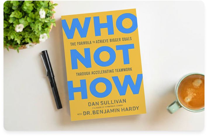 The book Who Not How sitting on a white desk with a black pen next to it.